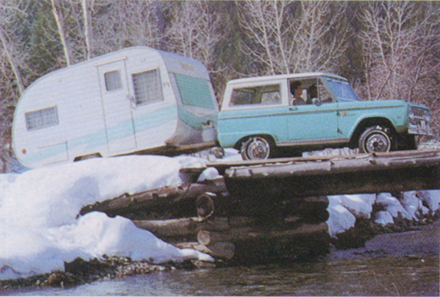 1967 Ford BroncoSUV towing a caravan across a makeshift log bridge in a snowy landscape