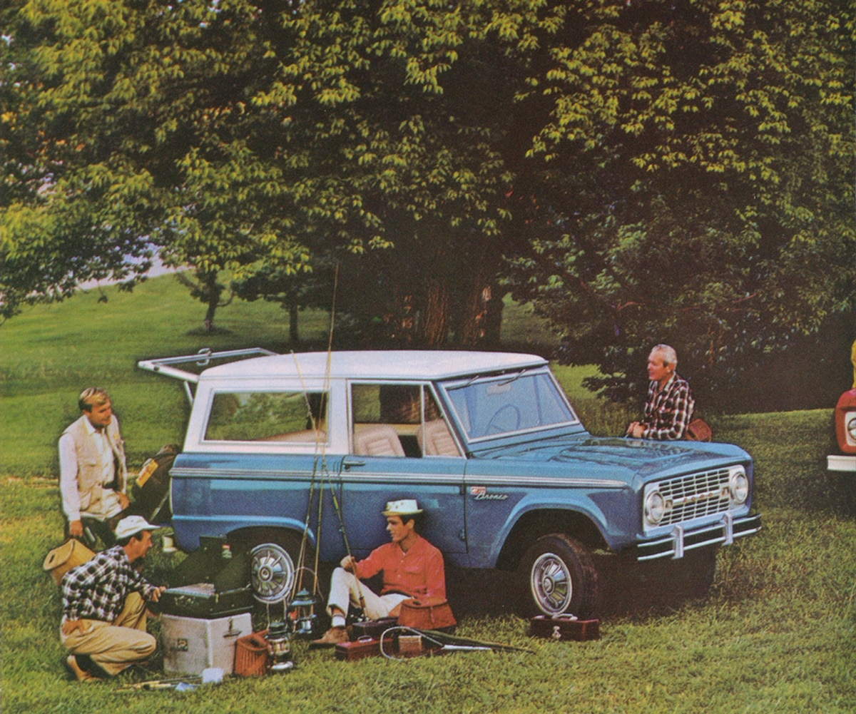 Light blue Bronco SUV with a white top parked in a grassy field with a group of people and camping equipment