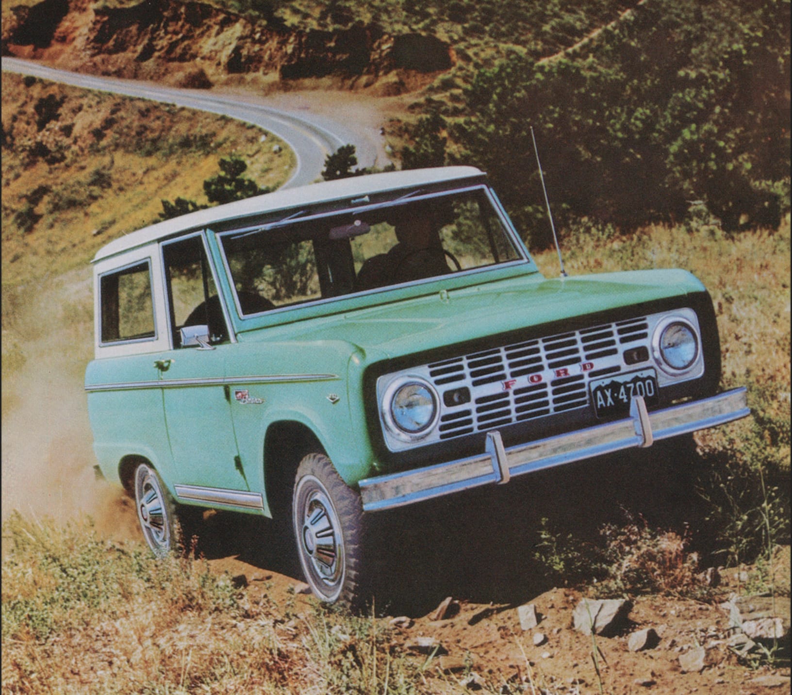 A turquoise 1967 Ford Bronco driving on a rugged dirt trail with a scenic winding road in the background